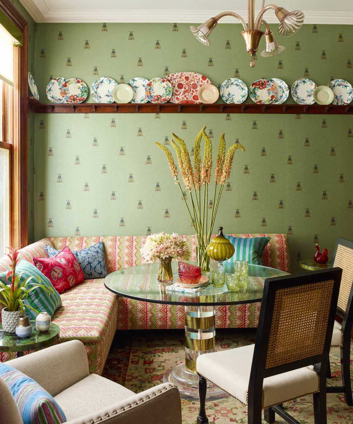 Green wallpapered dining nook with striped banquette seating, glass circular dining table and rattan backed cream chairs