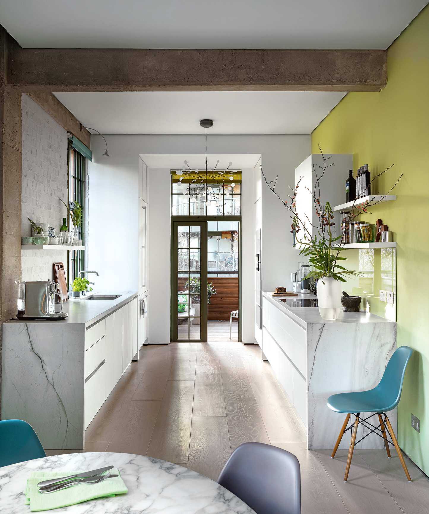 A marble galley kitchen with yellow wall, open shelving and teal chairs.