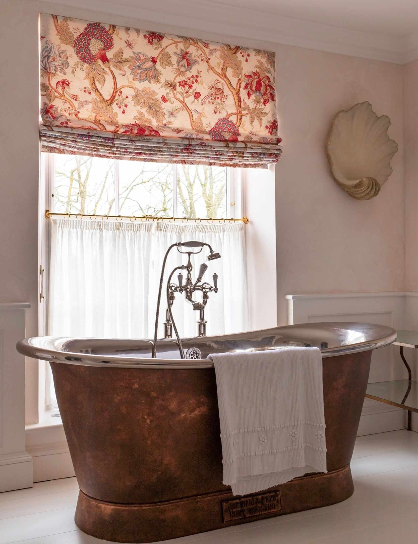 Double-dressed bathroom window area featuring a patterned roman shade layered above a panel of semi-sheer white cafe curtains.