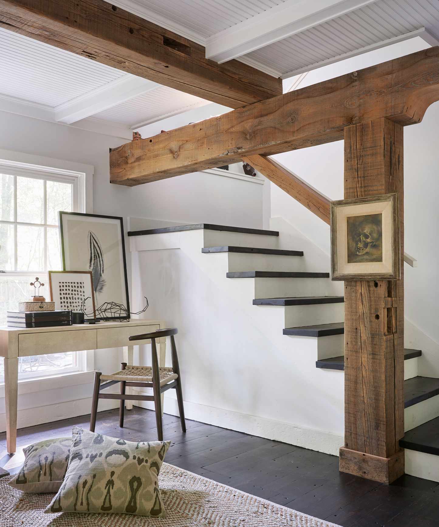Basement in period property with exposed wooden beams and columns, white wooden paneling, white and wooden stairs down into the basement, wooden desk facing window