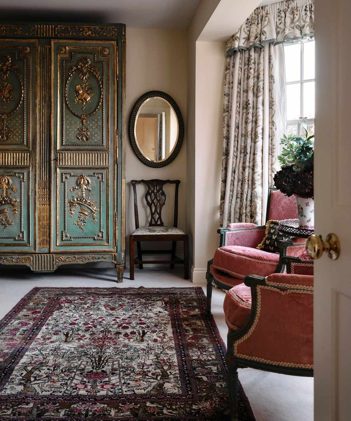 Traditional living room with pale pink walls, persian rug on floor, antique blue armoire, two pink antique armchairs, dark wooden mirror and large window with pelmet curtains