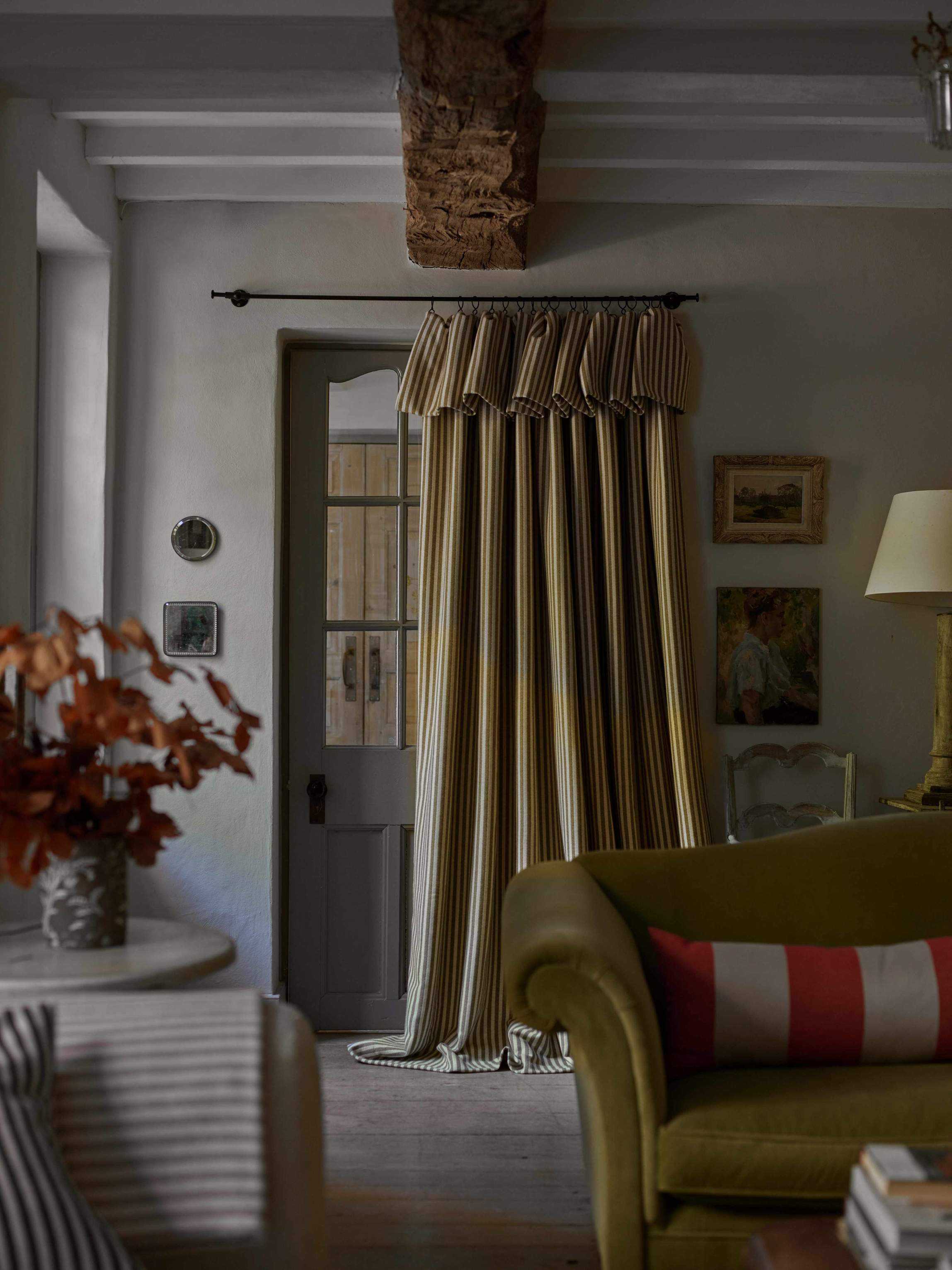 gray and green living room with stripe curtain in front of door, green couch, stripe cushions, beams, paintings, fall stems in vase on side table