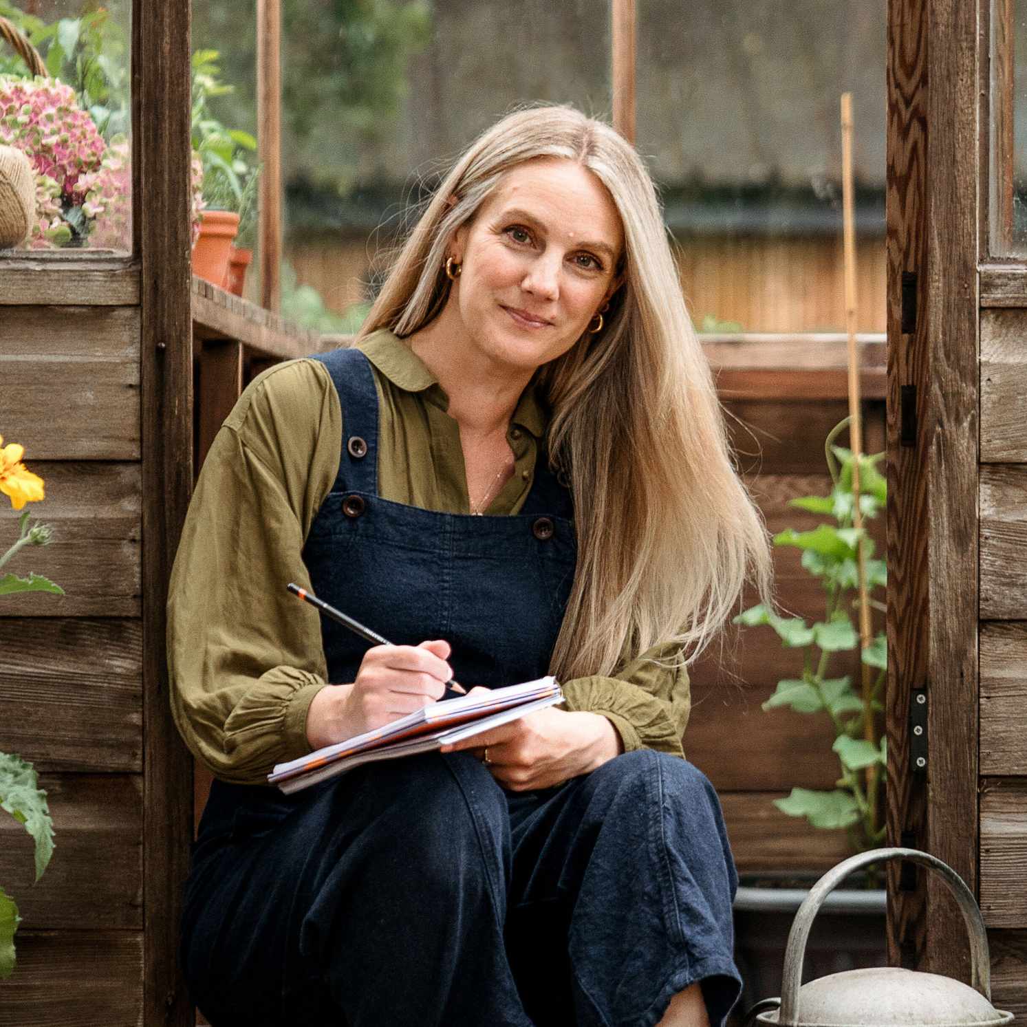 Woman sat on greenhouse step wearing navy dungarees and khaki shirt and holding a notebook