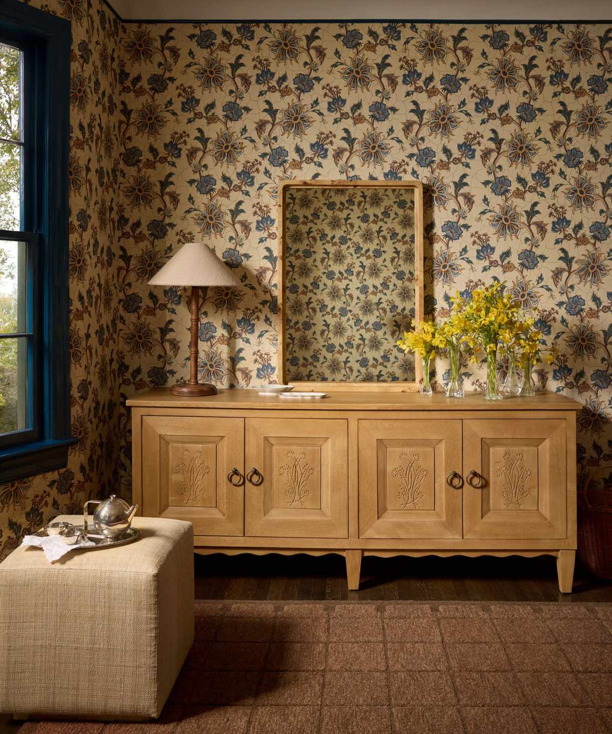 a room with a checker printed rug, a light wood credenza, a fabric ottoman, a mirror leaning on the credenza, a lamp, and an arrangement of flowers, all in a room with warm, floral wallpaper