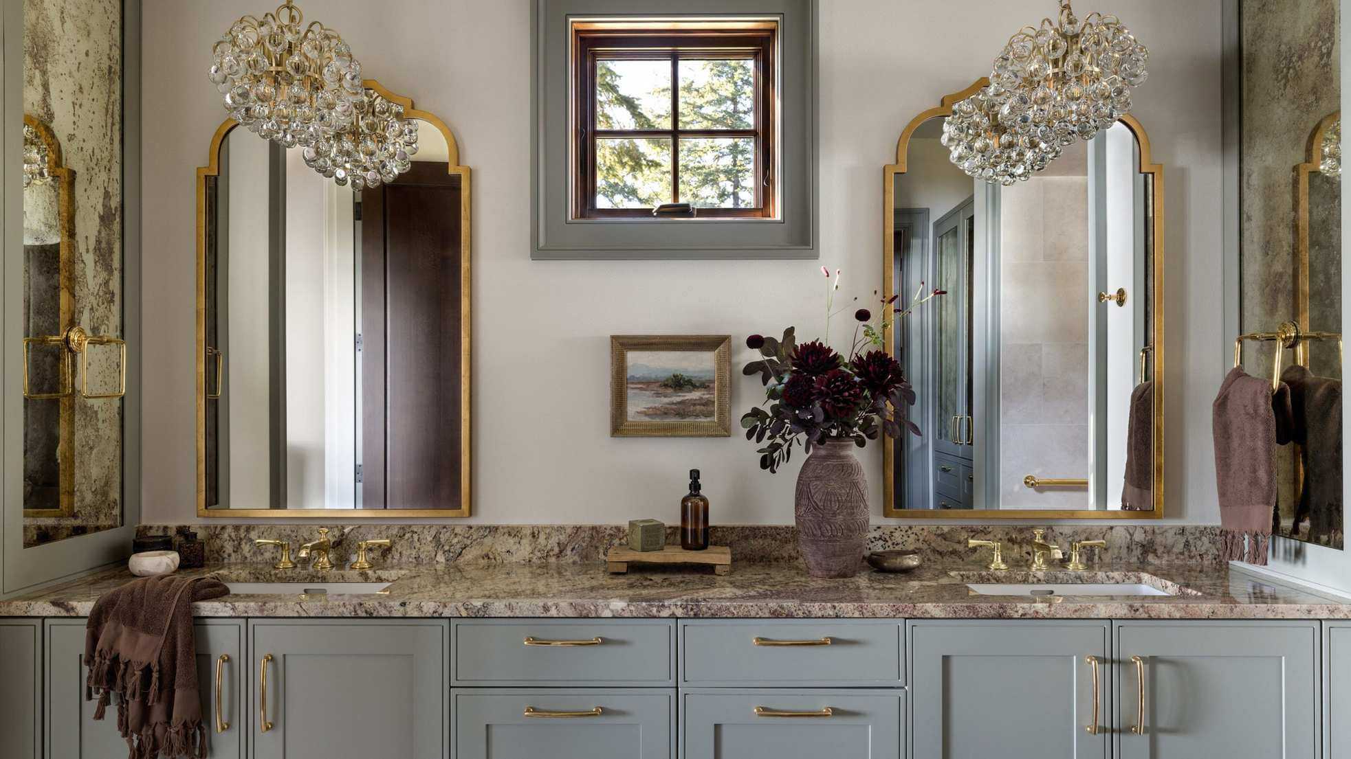 A sophisticated bathroom with warm white walls, pale blue cabinets, marble countertops, two brass mirrors on the wall, brass fixtures, and two glass pendant lights.