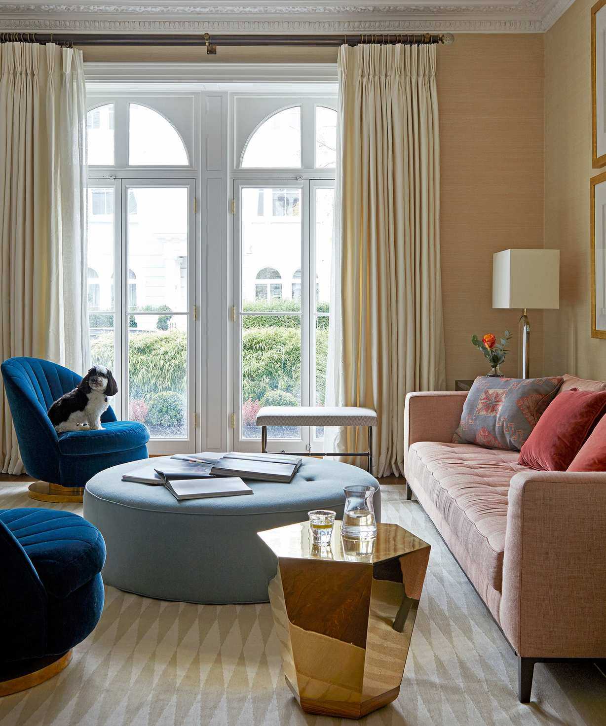 Living room with yellow textured wallpapered walls, cream diamond print rug, pink sofa with colorful cushions, two deep blue lounge chairs, golden metallic geometric side table, large rounded sky blue ottoman, large floor to ceiling window with cream curtains, cream and silver table lamp