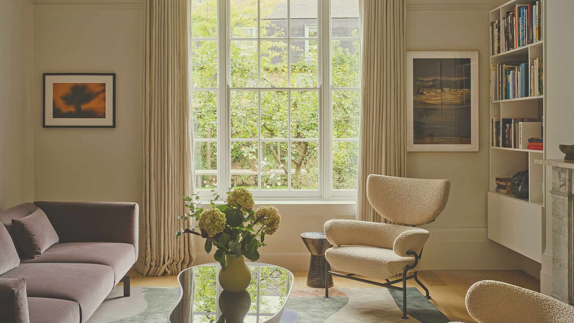 A sunlit contemporary living room by Atelier Como featuring a mauve velvet sofa, a cream boucl&eacute; lounge chair, and a large window overlooking a green garden.