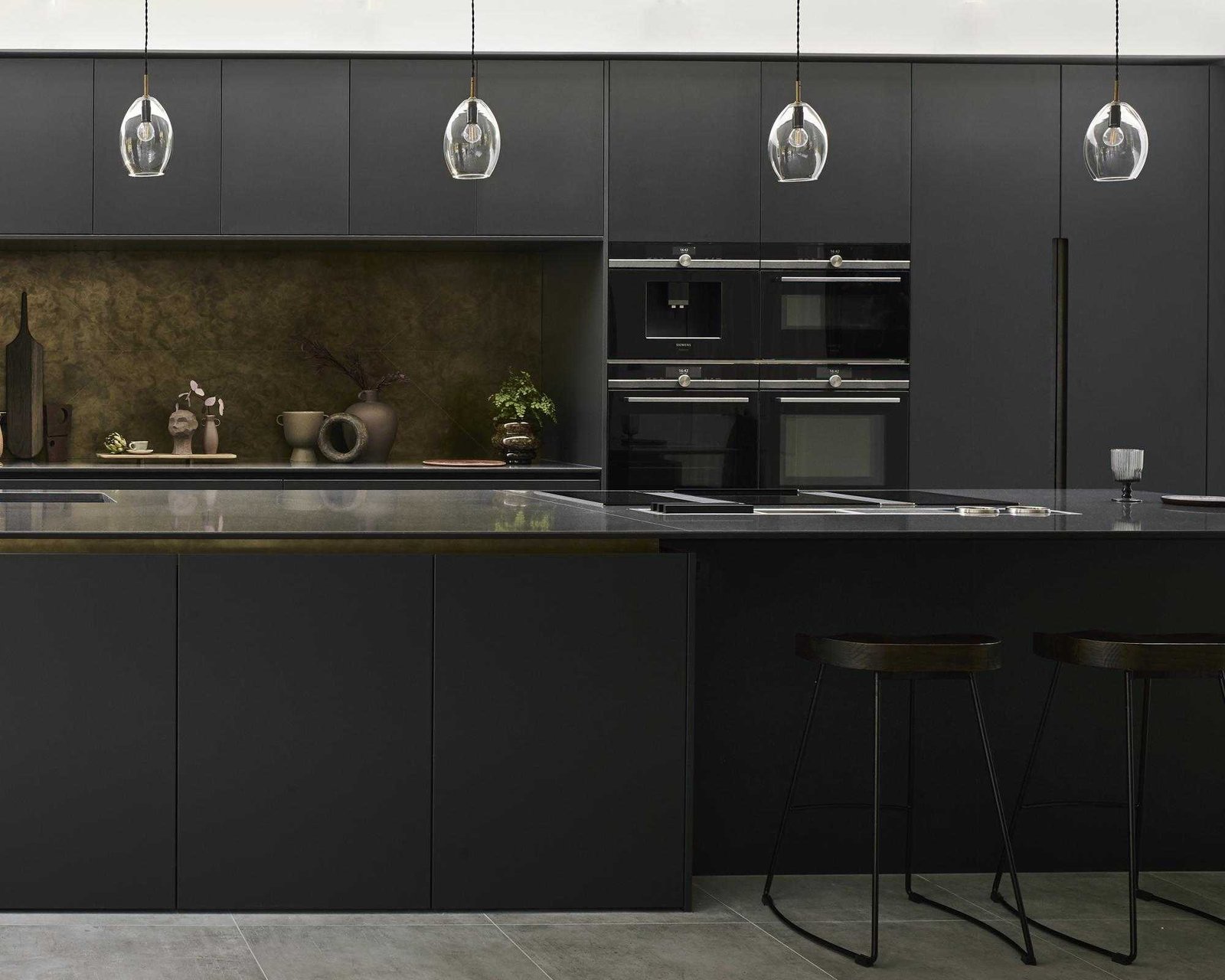 A black kitchen idea with matte black cabinets, stone worktop on the island, brass backsplash and pendant lights