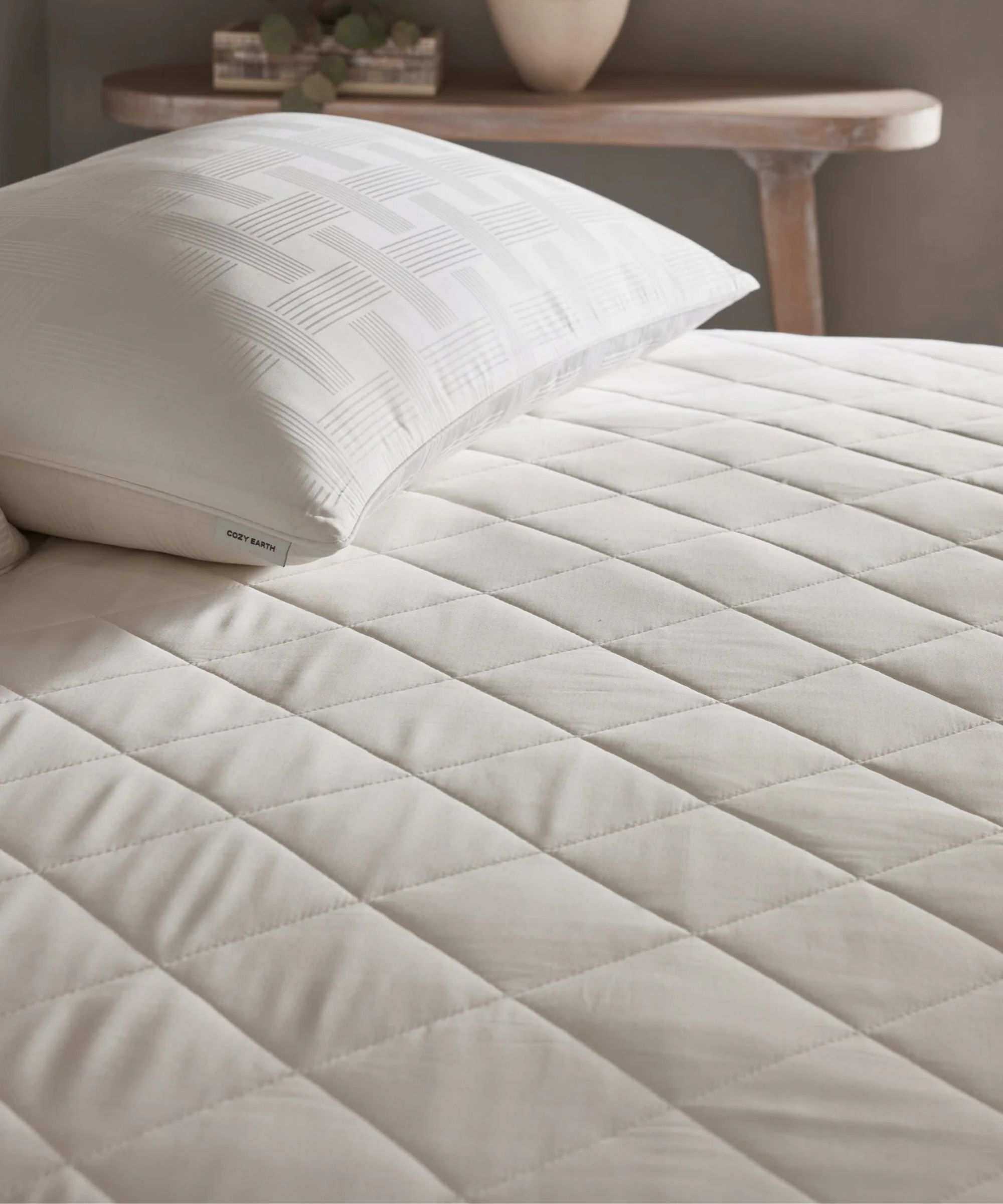 A white quilted Cozy Earth Bamboo Mattress Pad shown on a bed, with a white pillow. Behind is a wooden side-table, with a green houseplant in a brown rectangular pot, and a white plant pot beside it.