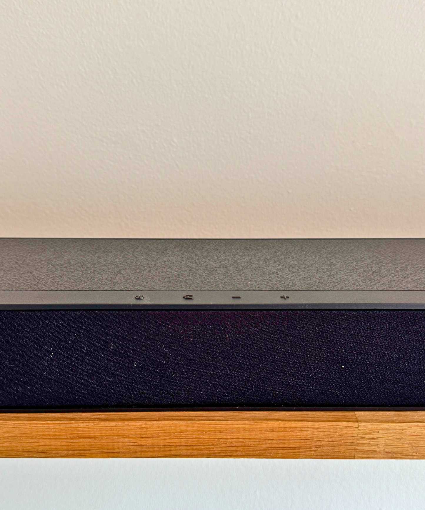 Closeup of Hisense HS2100 soundbar's four control buttons on wooden shelf in front of white wall.