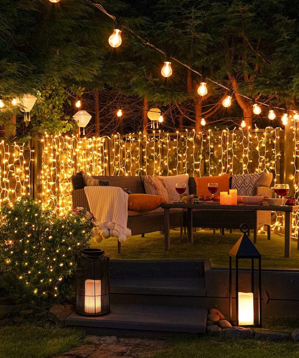outdoor sofa with cushions and blankets lit up by festoon lights, string lights and hurricane lanterns