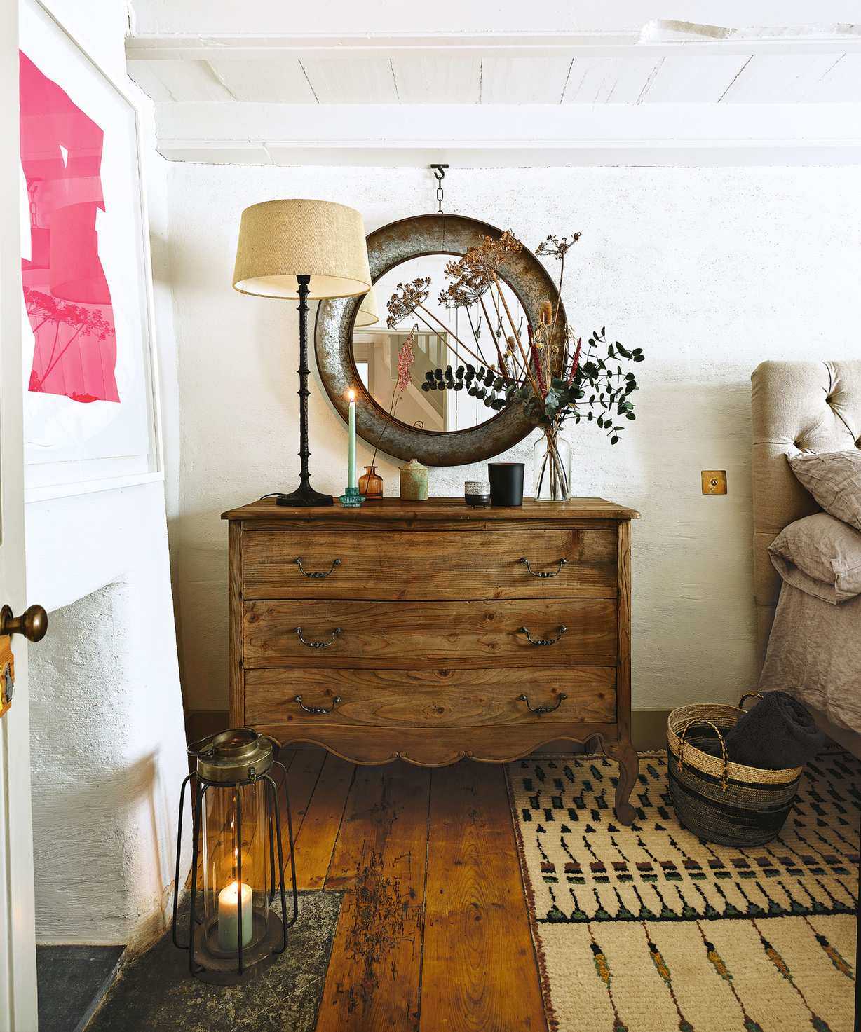 Cozy, country bedroom with textured cream walls, wooden chest of drawers, round mirror, table lamp, patterned textured rug, bed in corner