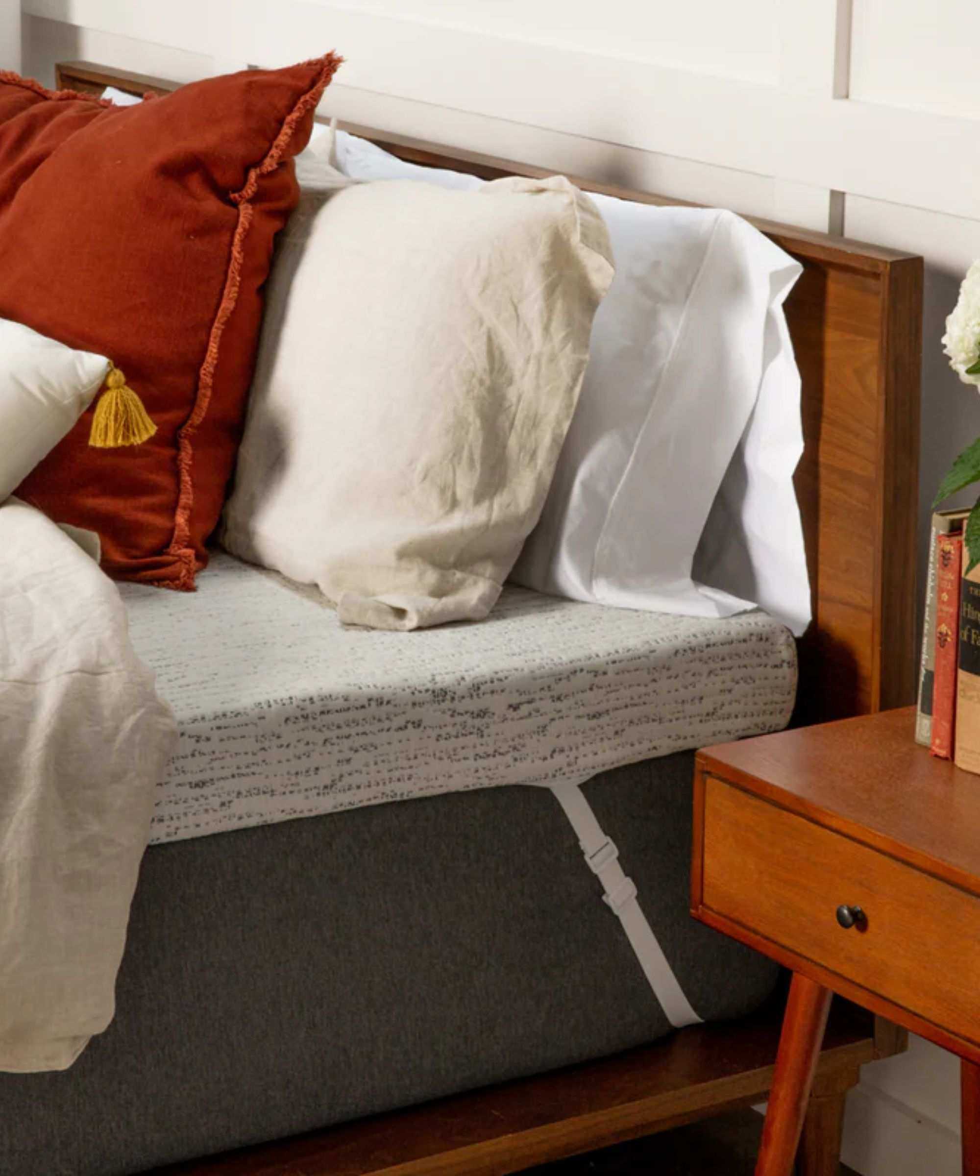 A grey Viscosoft High-Density Mattress Topper with corner straps a grey mattress on a wooden bedframe, with red, cream, and white pillows and cushions, against a white wall, beside a nightstand.