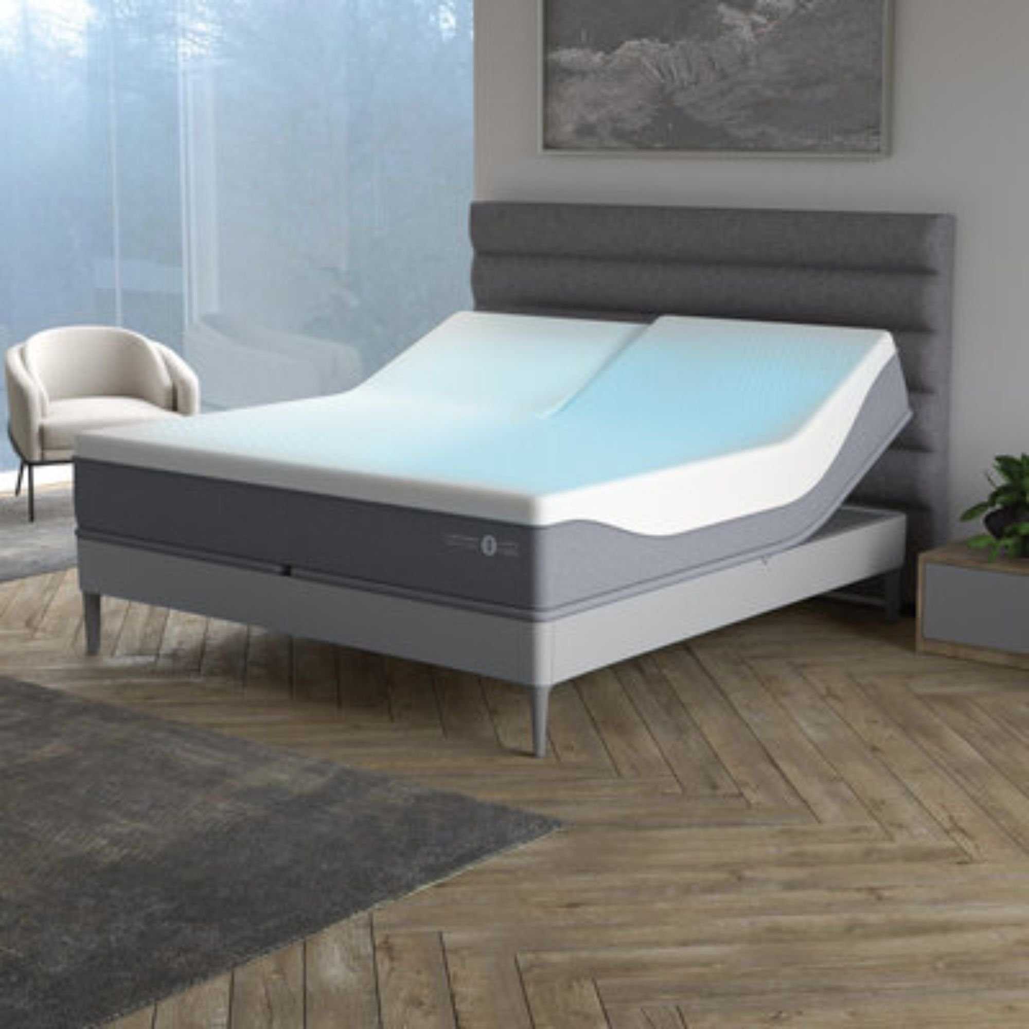 A grey fluted Sleep Number ClimateCool&reg; smart bed in a neutral bedroom with white walls, wooden flooring, a grey rug, large grey artwork above, and large windows to the left with a white boucle chair in front of it.