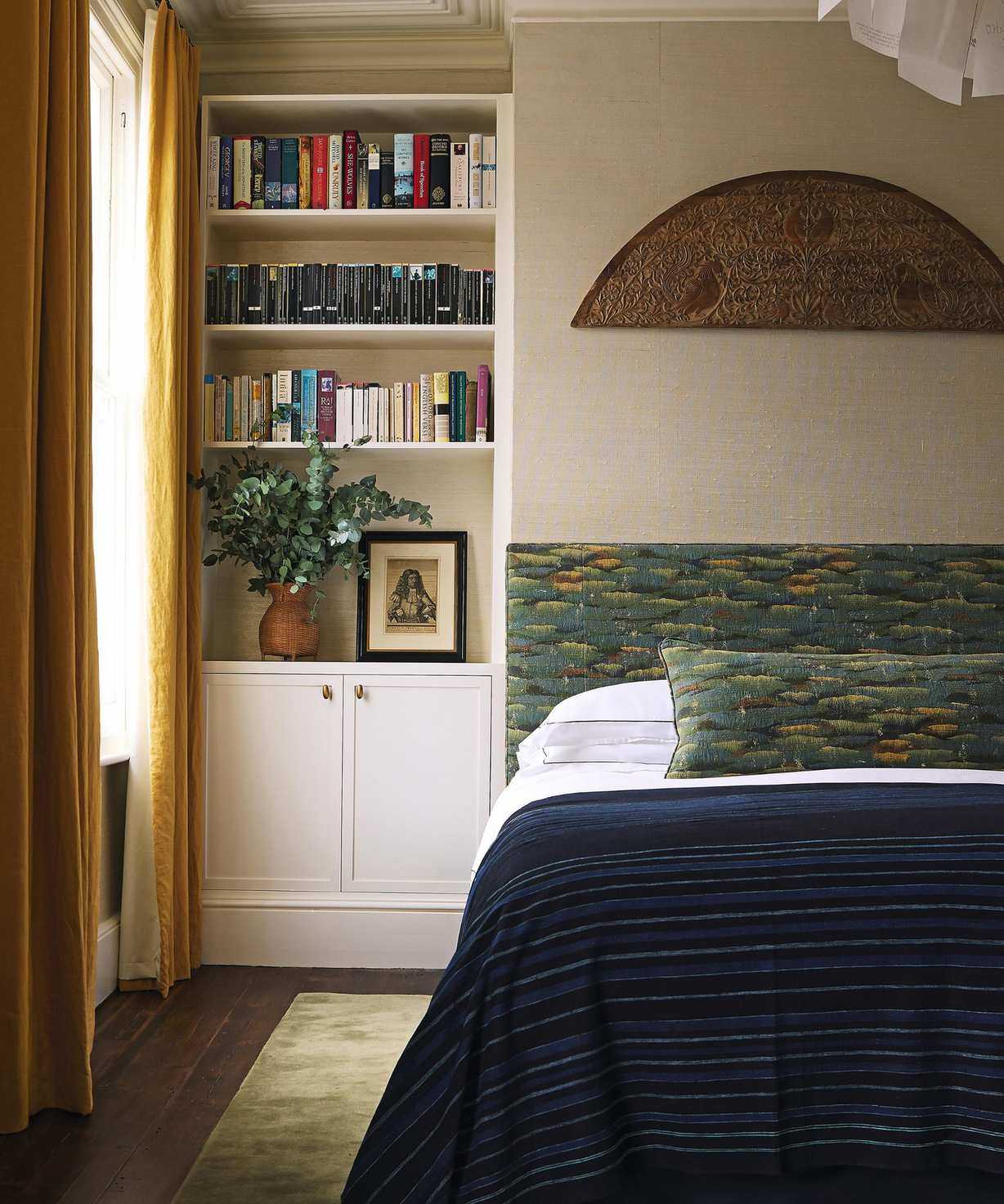 a neutral london bedroom with a bed place on the chimney breast, alcove bookcase and cupboard, yellow drapes at the window, and a green patterned custom upholstered headboarrd