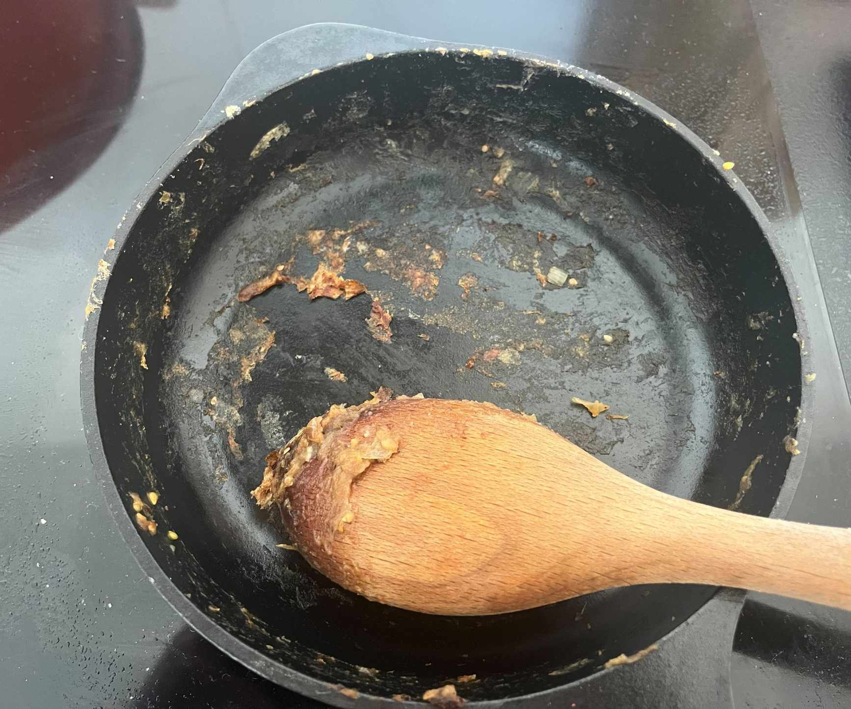 The stuck on remnants of food in a black cast iron skillet, with a wooden spoon.