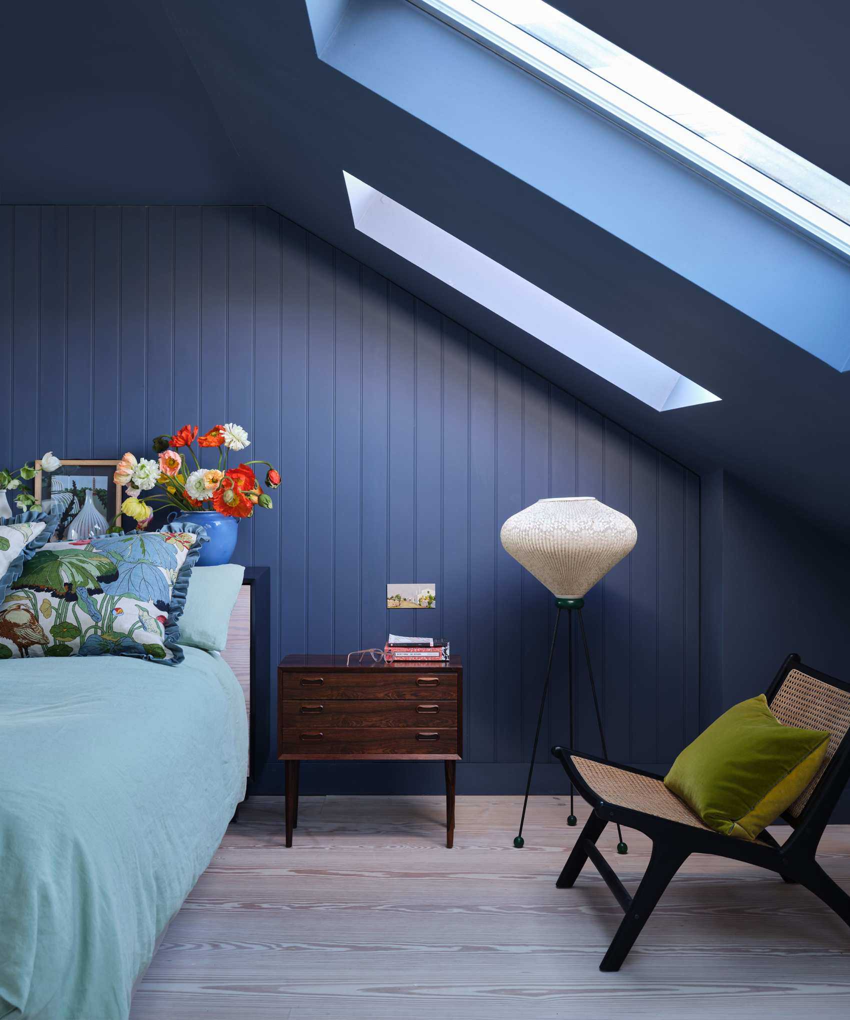 Color drenched bedroom painted deep blue, sloped ceiling with skylights, floor lamp, lounge chair, bedside table