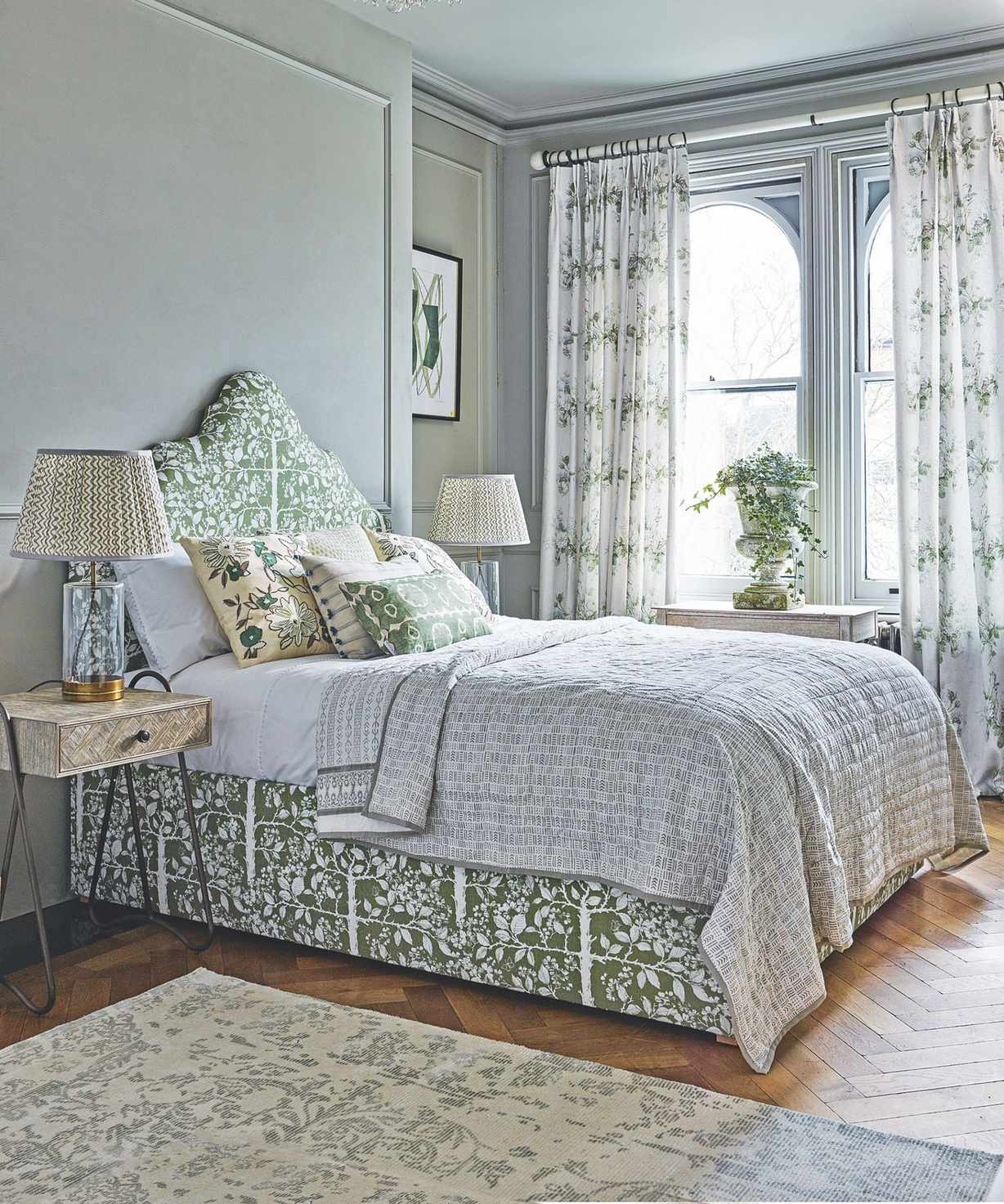 A pale green bedroom with windows, sage panelled walls, a green patterned double bed, patterned bedside tables, and patterned lamps. The floor is light wood and polished, with a large patterned rug.