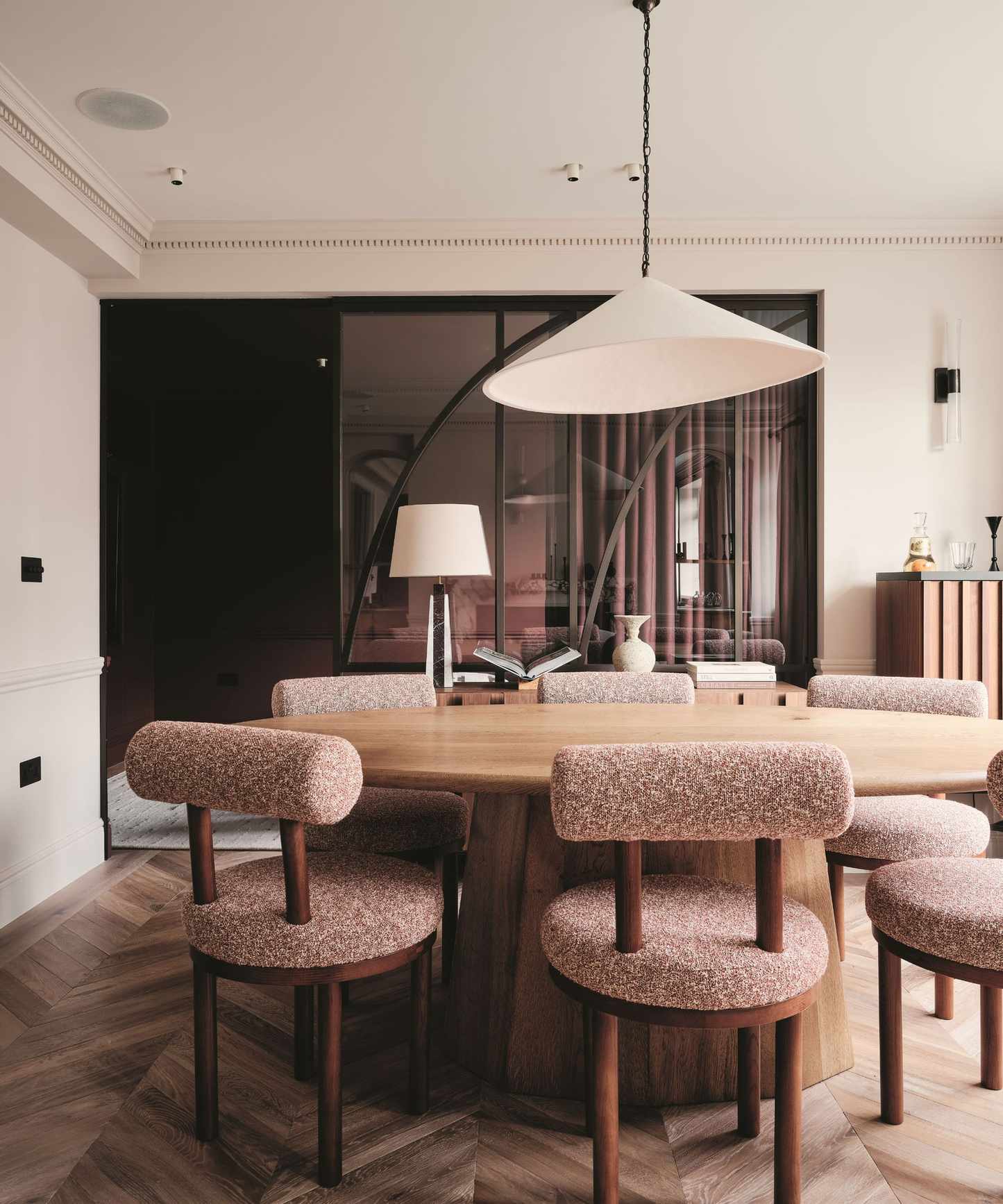 Oval oak dining table with mauve boucl&eacute; chairs under a large conical pendant. A curved glass partition and chevron wood floors complete the modern, warm interior.
