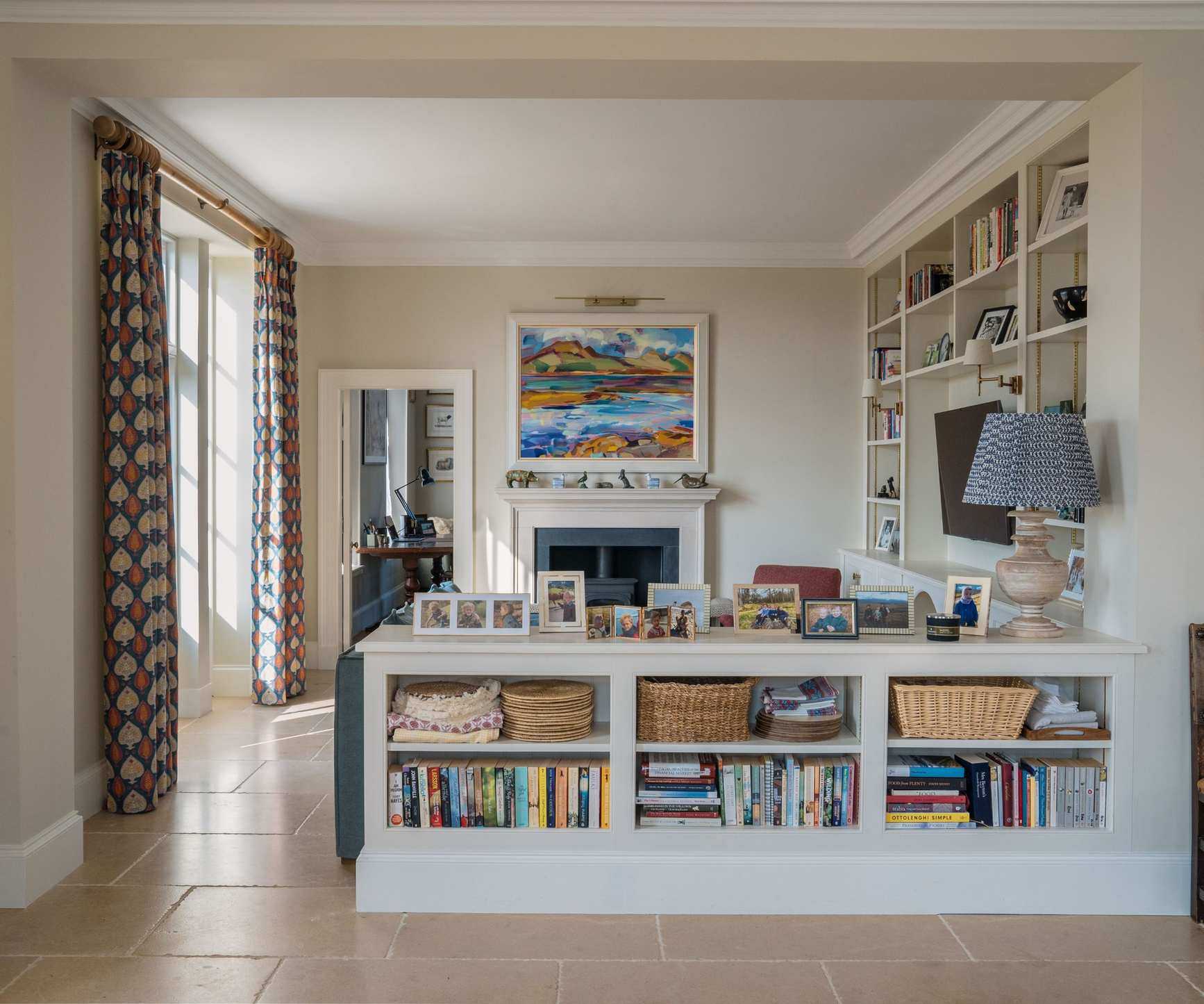 Living room with tiled floor, cream walls, French doors with floral drapes, a built-in storage unit with books, trinkets, and a TV at the centre, and a storage island