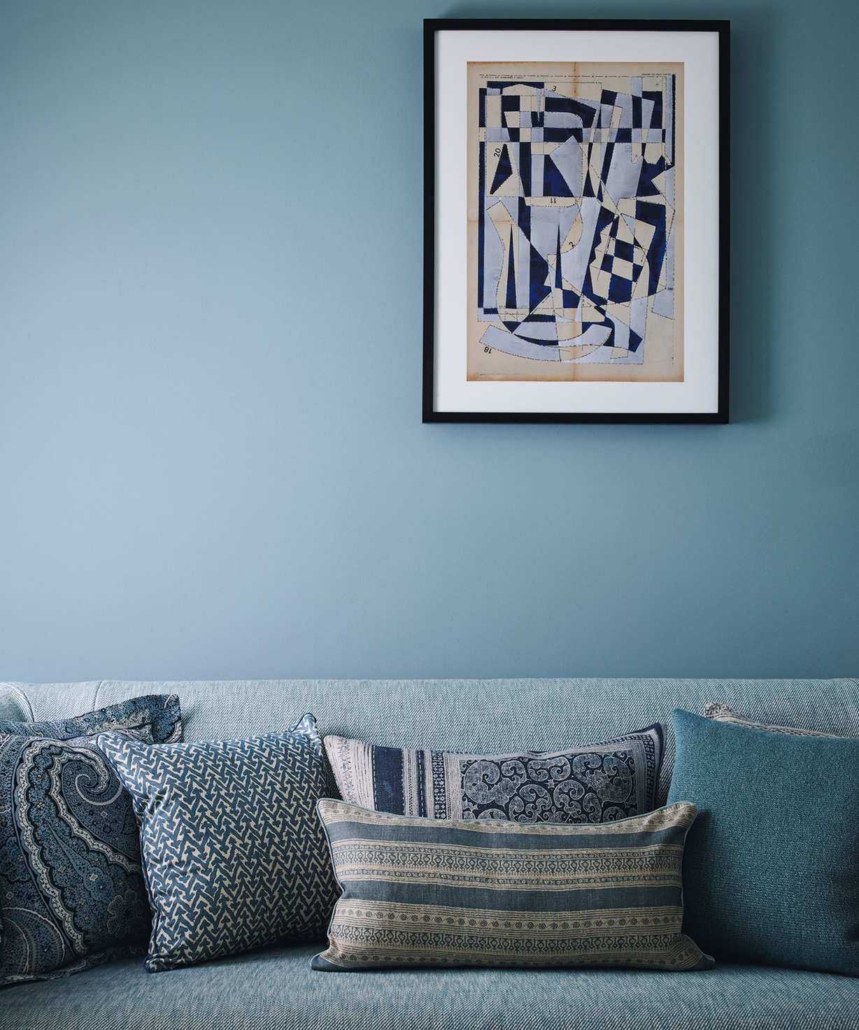 A blue sofa with assorted blue patterned cushions with a blue wall behind and a blue artwork in a black frame above