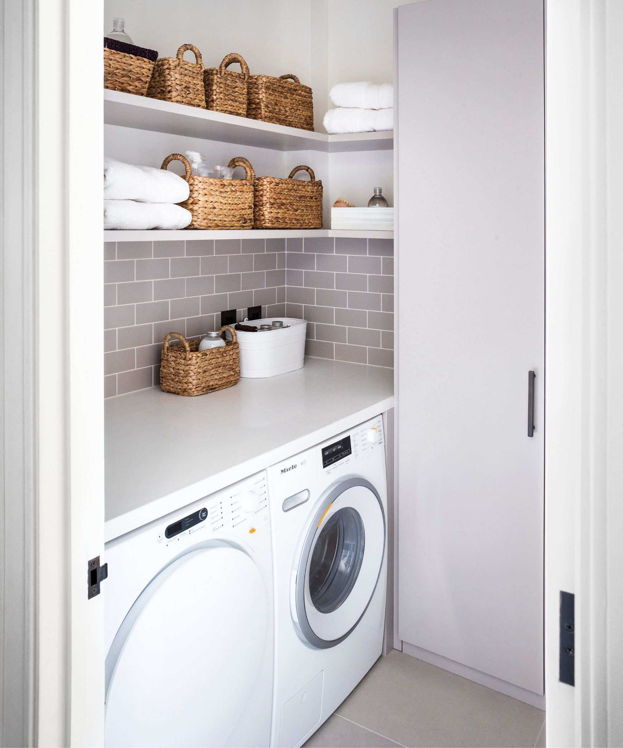 A clean and tidy purple tiled laundry room with a washing machine, tumble dryer, and shelves filled with wicker storage baskets and towels.