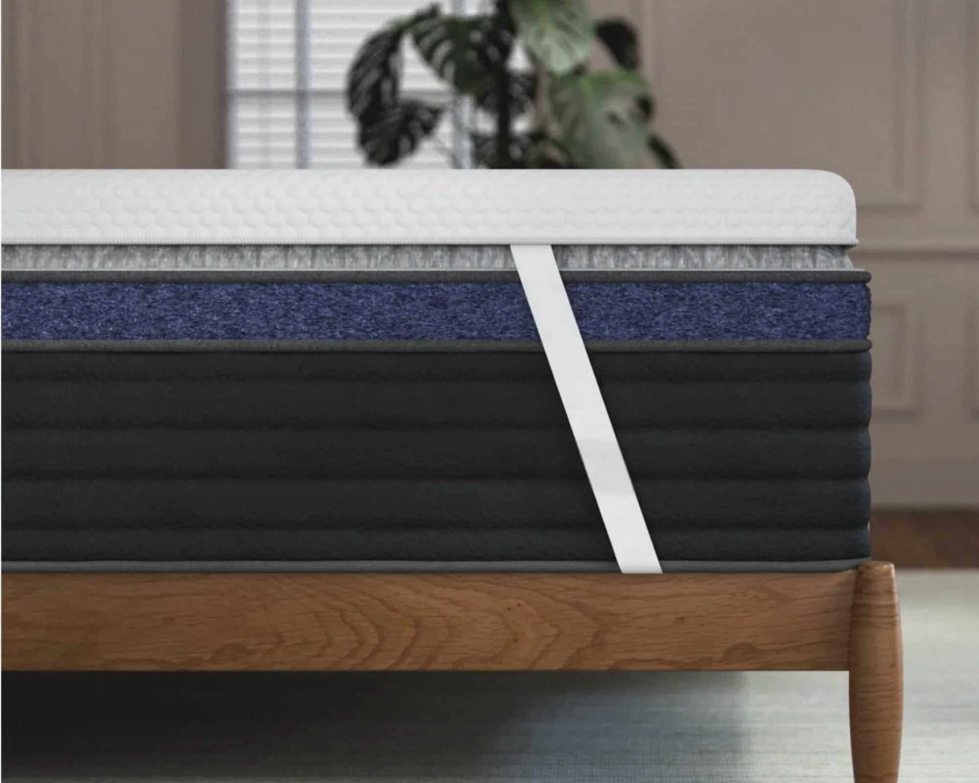 The white Helix ErgoAlign Mattress Topper with corner straps seen on a navy mattress and wooden bedframe. The floor is grey, and behind is a neutral panelled wall, cheeseplant and window with blinds.