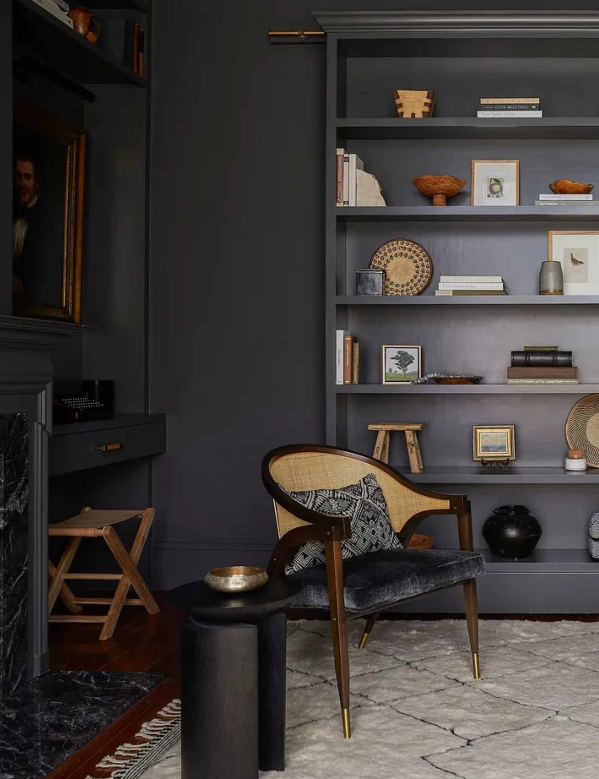 Tall, dark gray built-in bookshelf filled with books, baskets, and framed artwork, next to a cane-backed chair