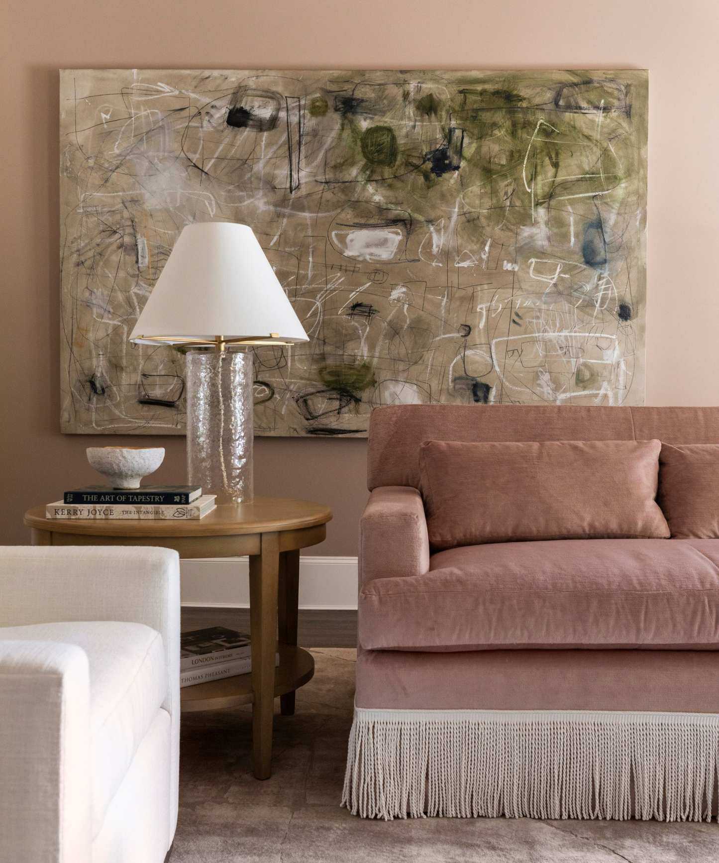 Pink living room with pink velvet fringed couch, large artwork, wooden side table and glass lamp