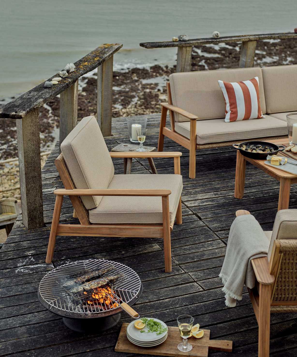 A wooden outdoor armchair with cream upholstered cushions on a wooden deck beside the sea. A small BBQ firepit beside it.