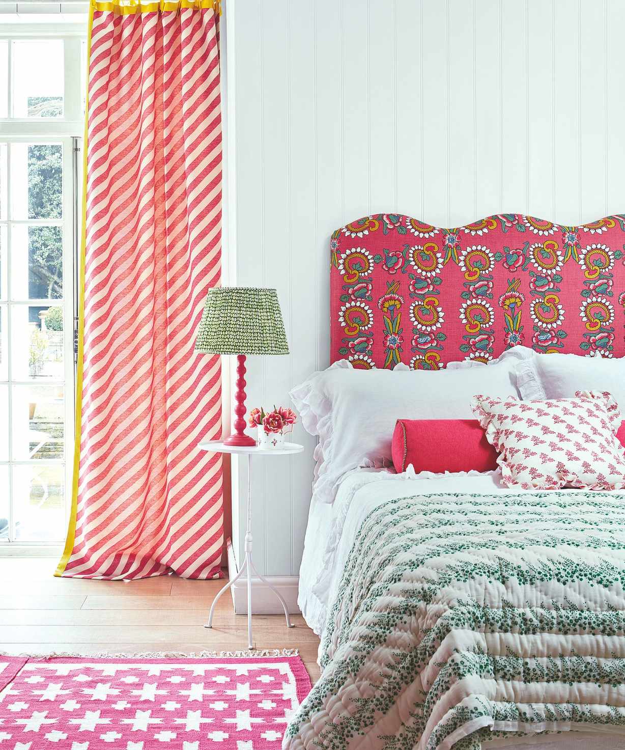 A colorful bedroom with white panelled walls, wood floors, pink patterned headboard, cushions and rug, white bedding, and green bedspread. Large window with red and white striped curtains on the left.