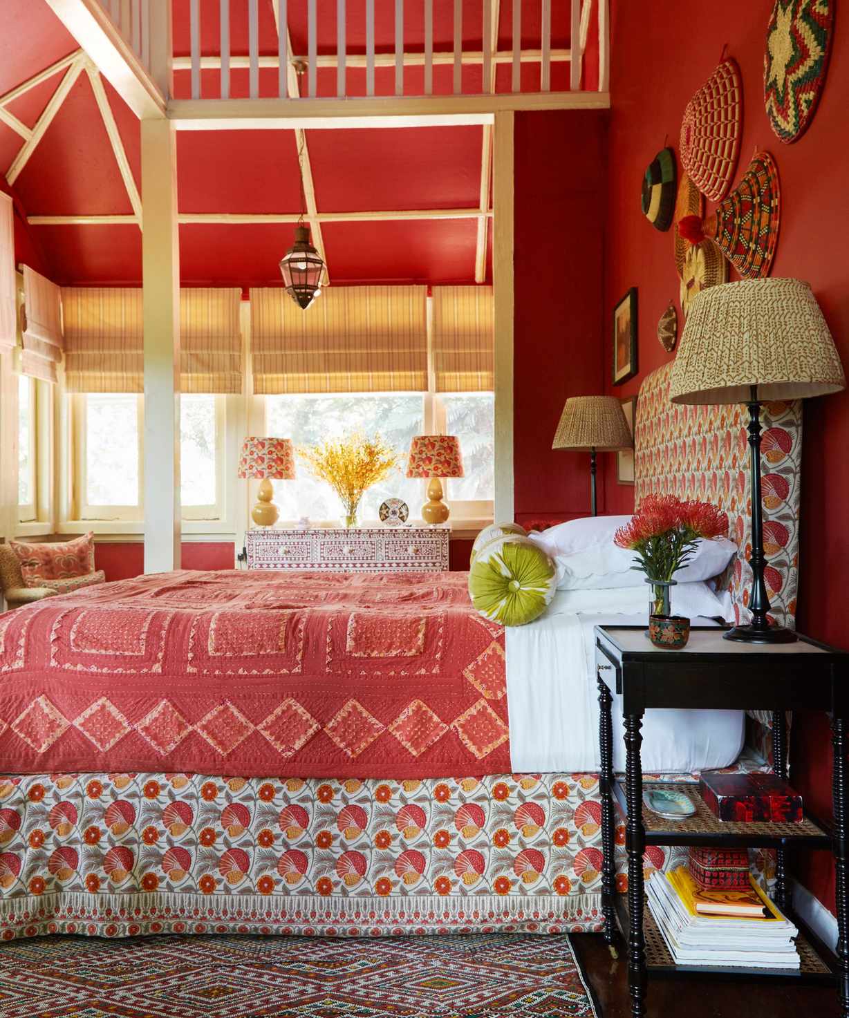 A side-on view of a bright red bedroom, with a large bed with floral-patterned bedding in white and red, two matching lamps on either side, a black bedside table with a stack of books.
