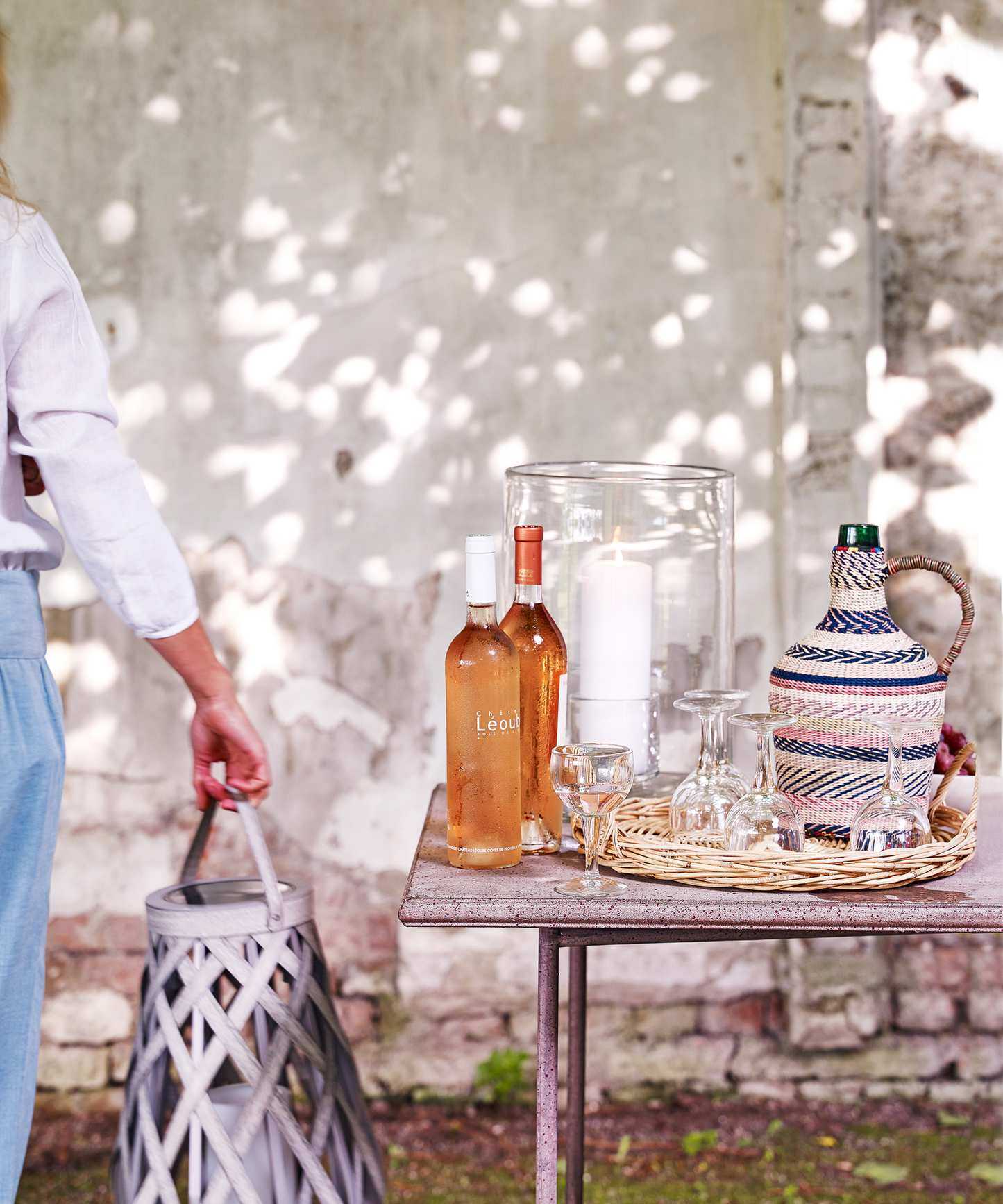 An example of what to include in garden bar ideas, with two bottles of rose wine, a large woven jug and wine glasses on a wicker tray in front of a white painted brick wall.