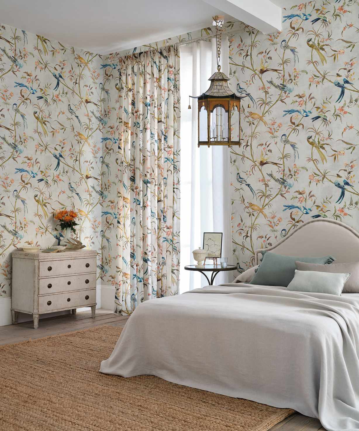 Large bedroom with matching floral patterned curtains and wallpaper, bed with upholstered headboard, hanging lantern light, natural textured rug