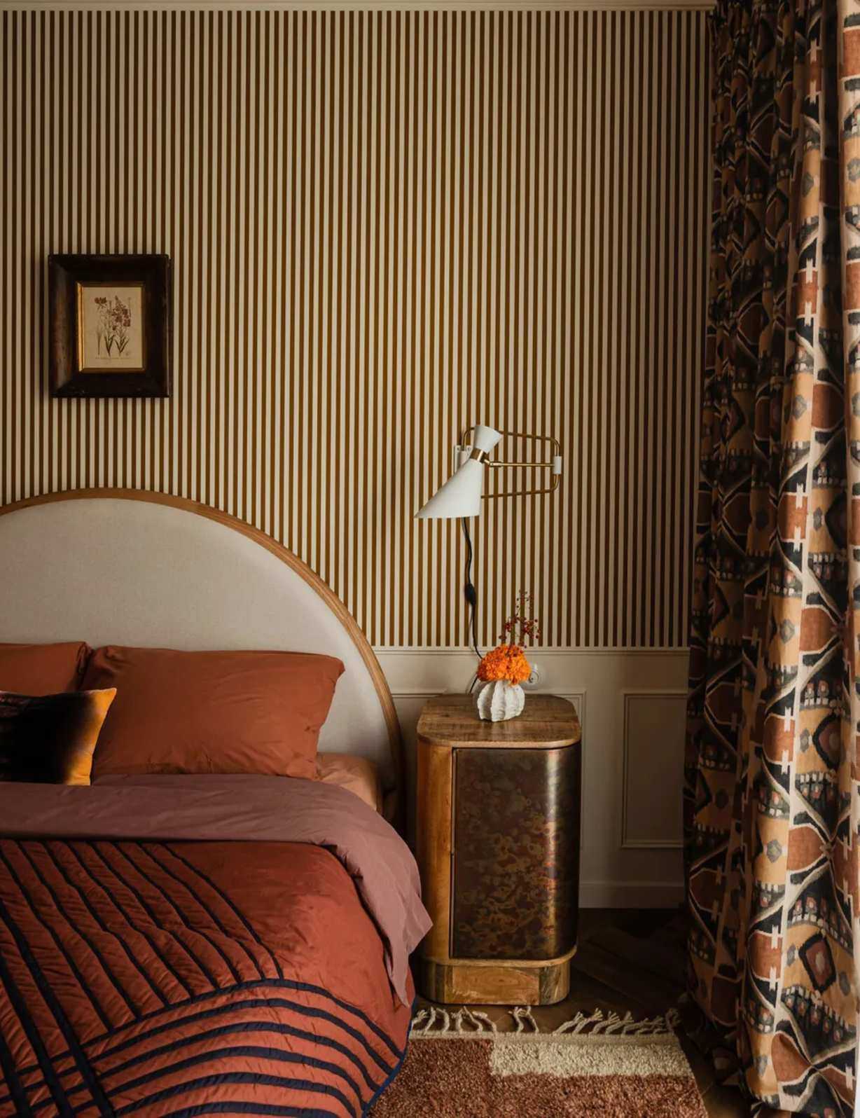 Art Deco style bedroom with striped wallpaper, curved headboard and orange bedding, geometric curtains and burl wood and gold bedside table