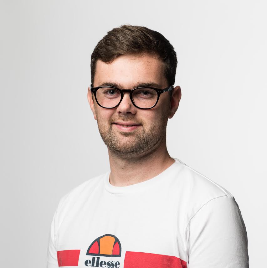 Luke Stitson wearing white and red t-shirt and glasses in front of gray background.