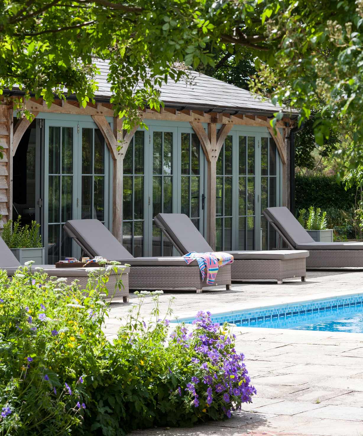A Mediterranean style garden room surrounded by plants next to a pool area with sun loungers