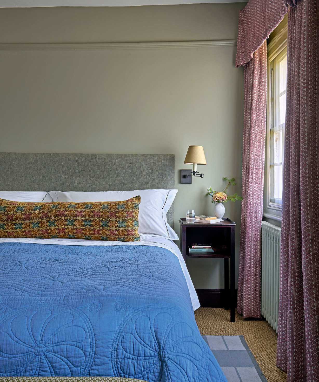 Bedroom with sage green walls, grey fabric headboard, blue quilt and long colorful bolster, rattan carpet and pink pelmet curtains around the window