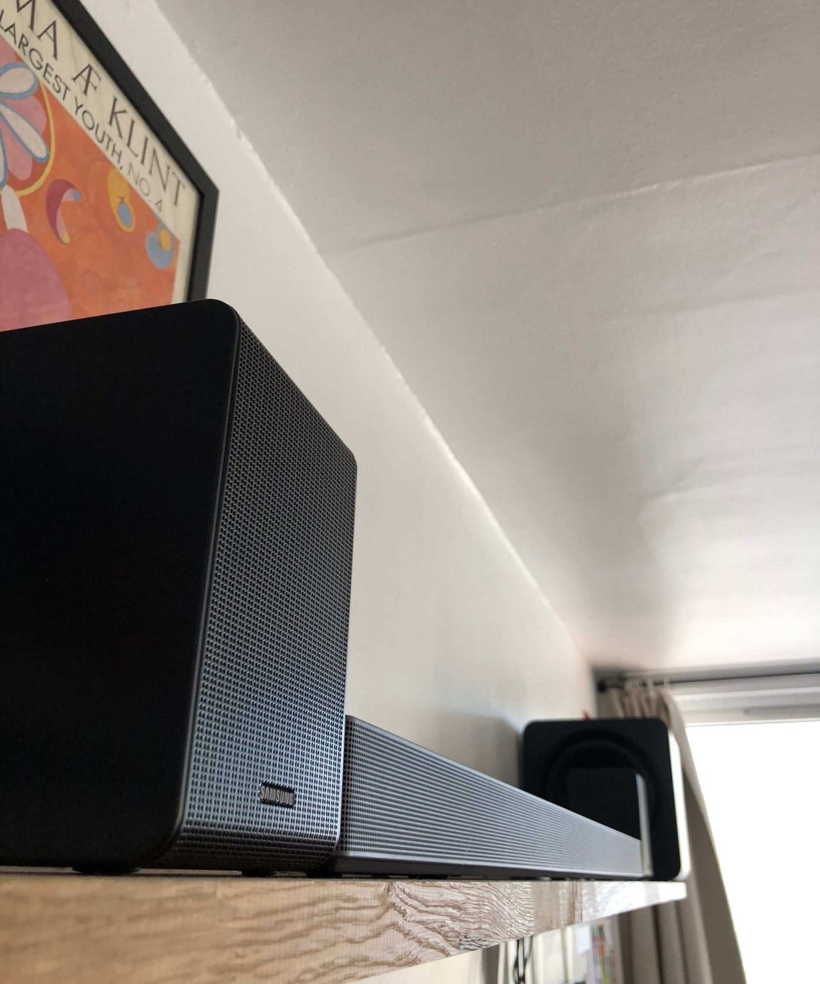 Samsung HW-QS750F soundbar on wooden shelf in Dan Fauzi's home, with curtains and bright window in background and art print pictured to the left.