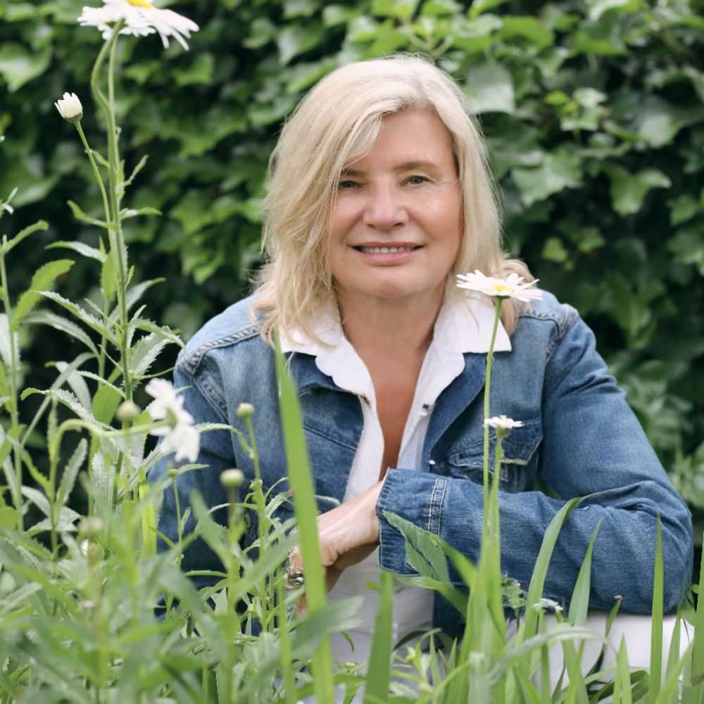 A headshot of Alicia Sokowlowski wearing a white shirt and blue denim jacket sitting behind grass and daisies with a green leafy background