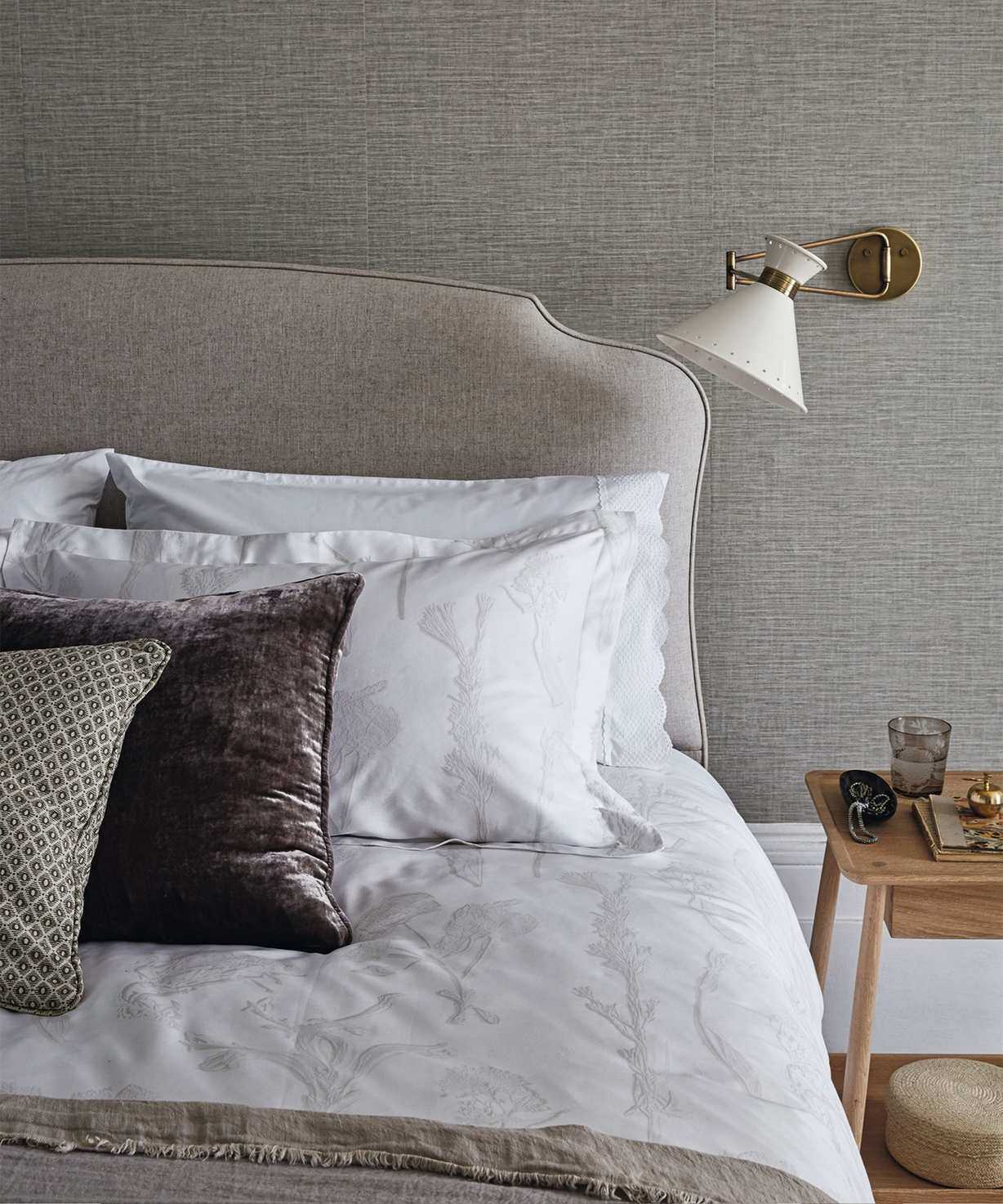 A neutral bedroom space with taupe walls, a grey brown bed with white bedding, assorted cushions, and a wooden bedside table. Behind are white painted baseboards, and the flooring is hardwood.