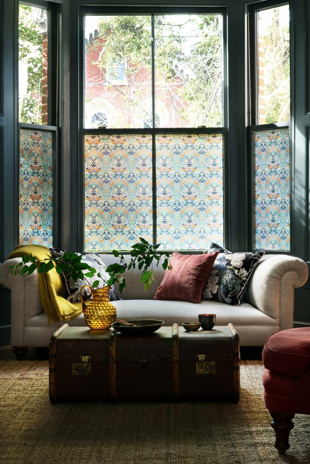 living room with green painted bay window, window film on windows, cream couch, floral cushions, amber glass jug, yellow blanket, vintage chest as a coffee table, rug