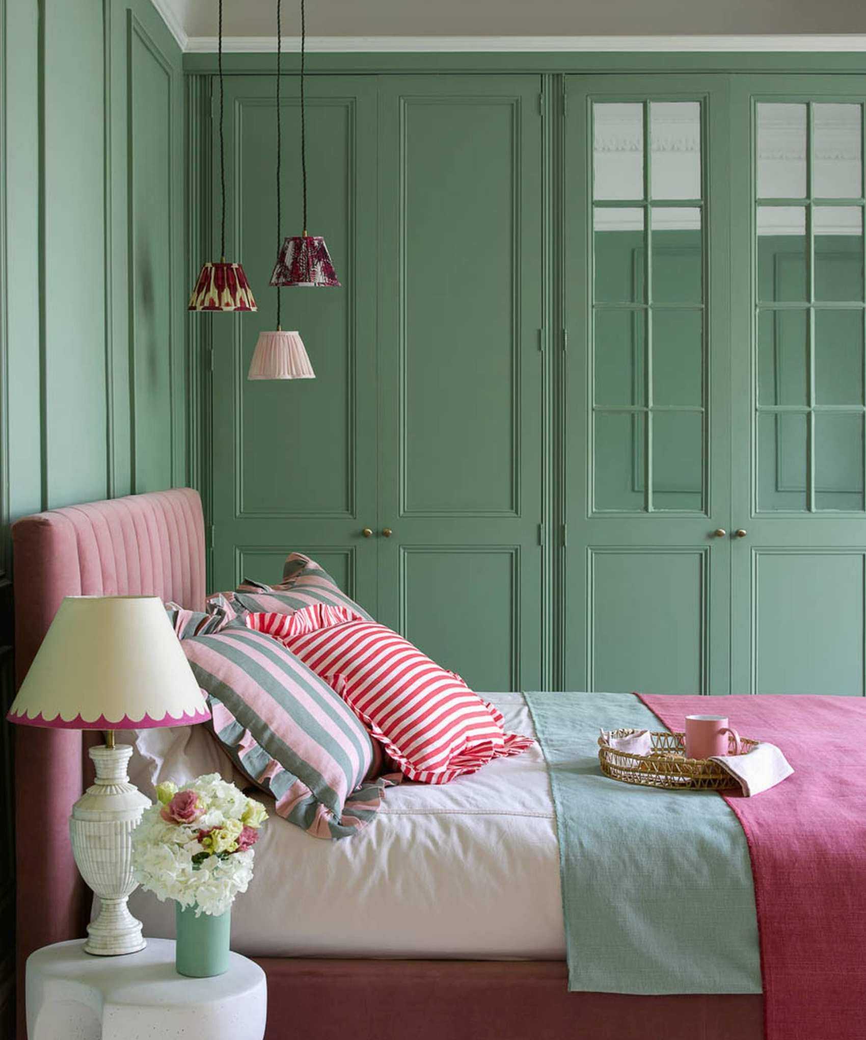 A colourful bed with a white bedside table and a lamp on it as well as a bunch of flowers. There are wardrobes painted in green