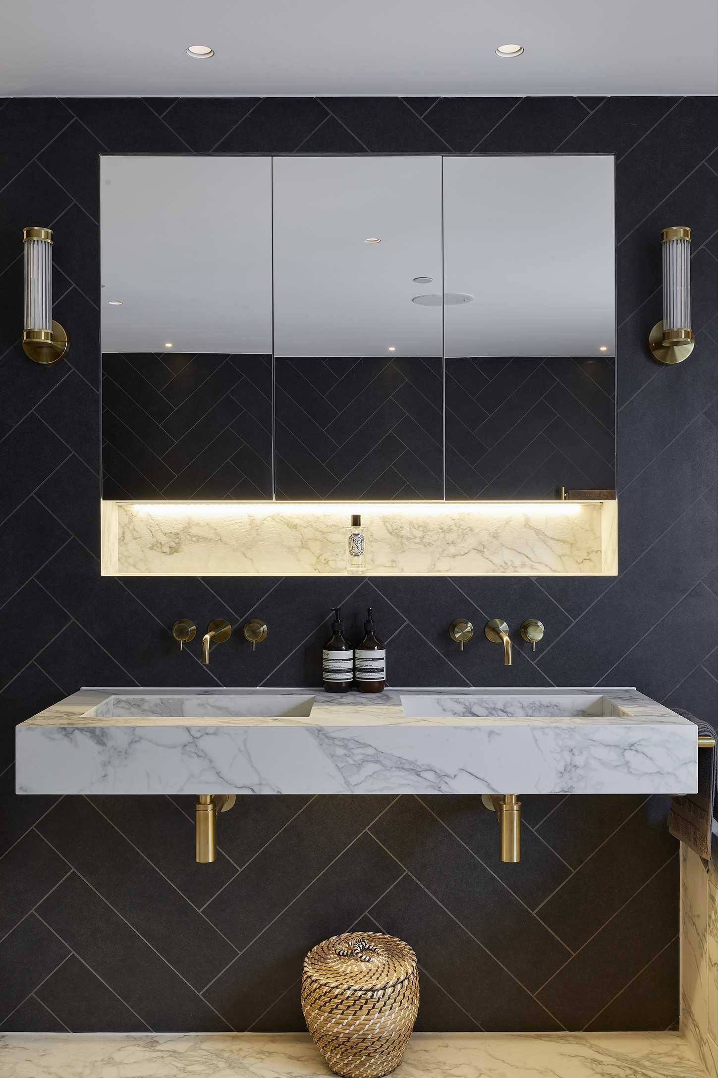 An example of dark bathroom ideas showing a marble basin with gold taps, gold wall lights and a mirror, and navy herringbone tiles