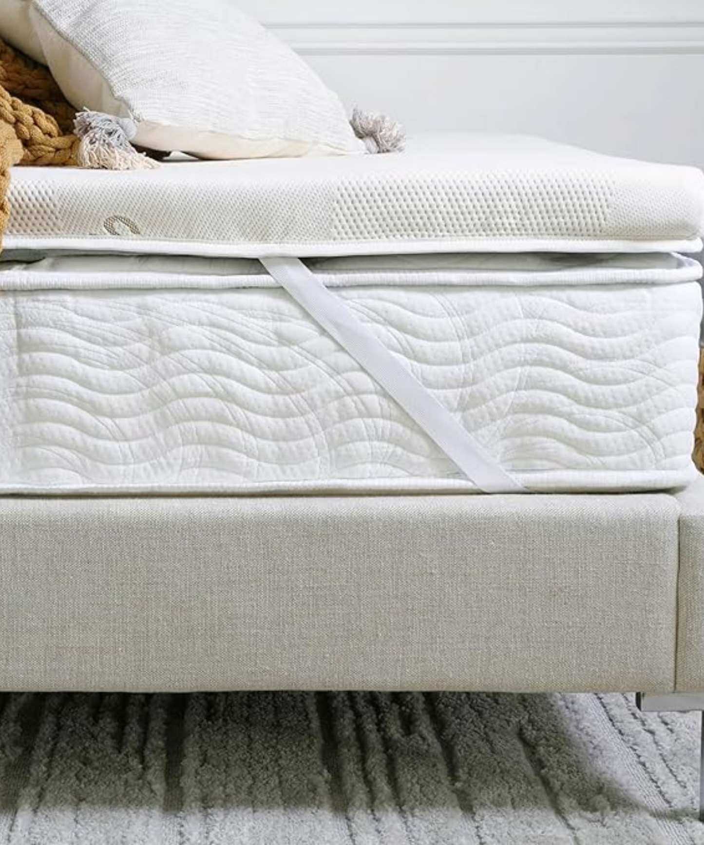 The Saatva Graphite Mattress Topper on a white mattress on a neutral bedframe, with a white cushion and an ochre knitted blanket on top. The wall is white, and the rug beneath is grey and striped.