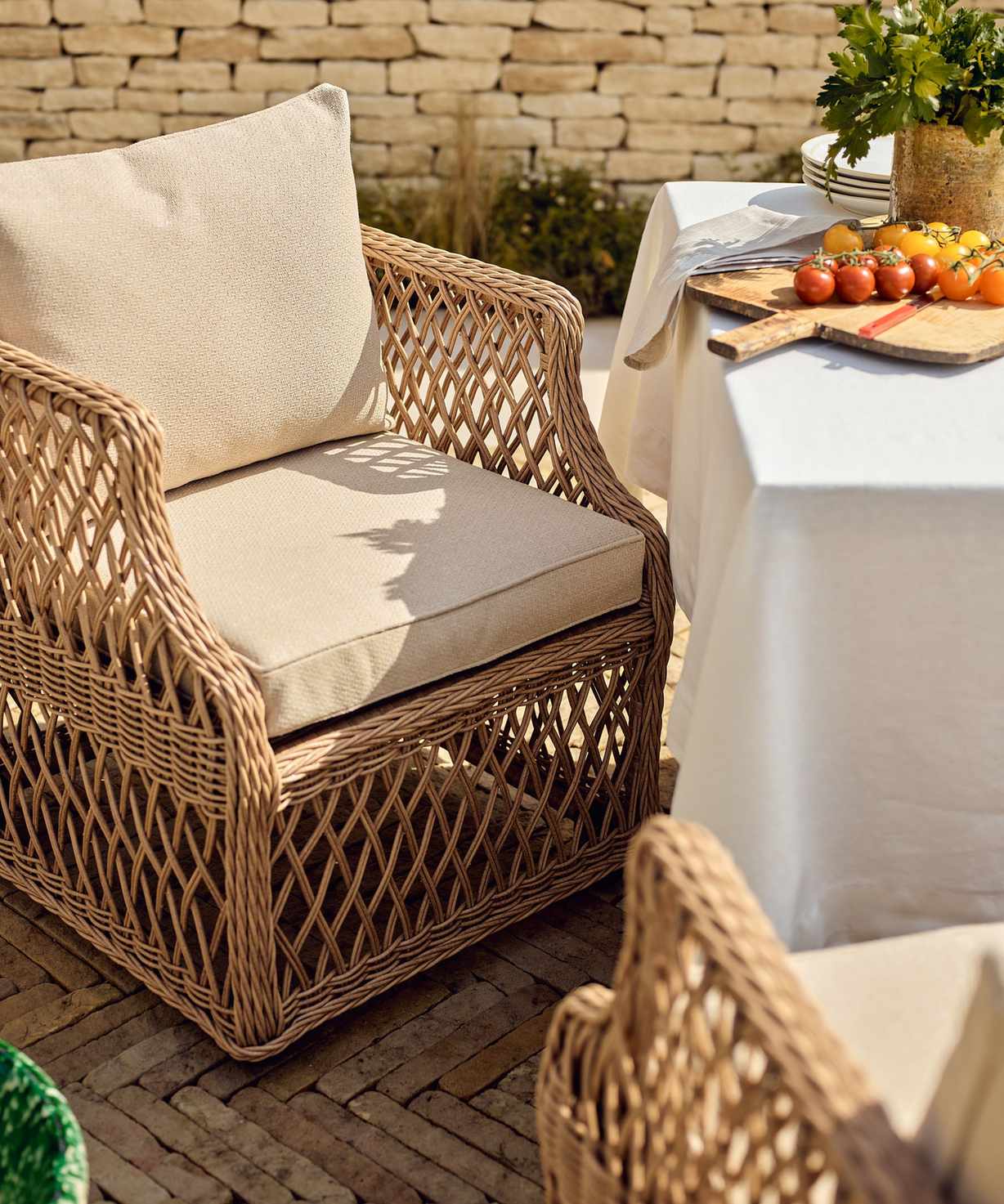 A wooden wicker outdoor armchair with cream upholstered cushions beside a garden table covered with a white cloth.